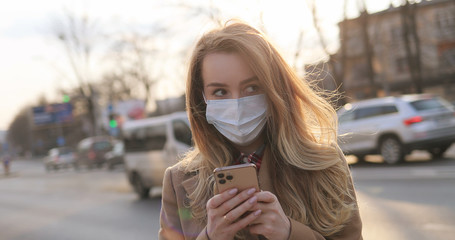 Caucasian young woman in medical mask texting message and tapping on mobile phone at street with road and cars. Girl typing and scrolling on smartphone in city outdoors. Pandemic healthcare concept.