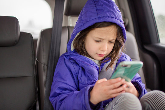 A Little Girl Plays With Phone In The Backseat Of A Car On A Car Trip