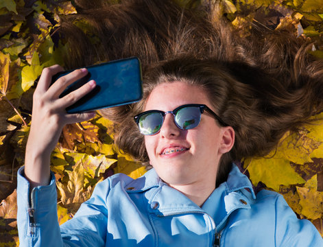 teenager girl wearing sunglasses and lying down on the floor covered by yellow leaves while taking a photo with her mobile phone to herself. The girl is smiling to the camera. Horizontal phot