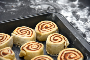 A pan of raw, homemade cinnamon buns ready to be baked, with white flour on black countertop