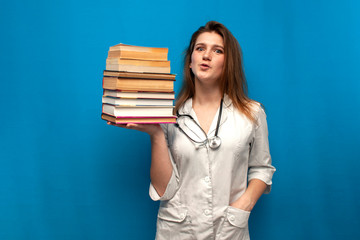 young beautiful funny girl the nurse holds many books on a blue background and is surprised, the student is studying in a medical college