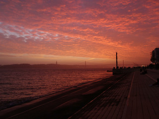 Naklejka na ściany i meble Sunset in Lisbon with the red clouds and the Tagus River, in the background the bridge April 25 and Christ the King and tourists.