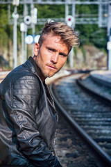 Obraz premium Attractive blond young man standing by railroad tracks, wearing black leather jacket and looking at camera