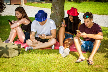 Group of young people sitting under tree, using their smartphones and communicating with each other or absorbed with them. Concepts of lack of communication, self-absorbtion, overuse of technology