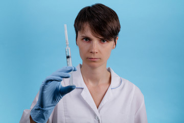 Pretty female doctor wearing white medical robe holding syringe at blue background. Young woman in medical uniforms and protective mask prepares a syringe. treatment and prevention of coronavirus