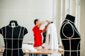 Black mannequins for sewing women's clothing are in the white room of the atelier for sewing clothes. A young man in red clothes is throwing a white dress on a mannequin.