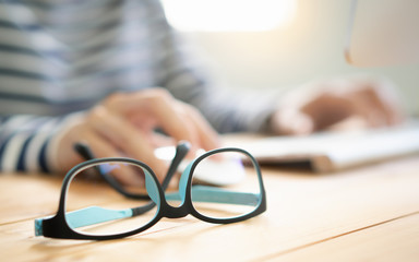 Closeup, Beautiful eyeglasses on wooden desk, hands of woman working with computer in background for long period of time which causes dry eyes, eyestrain and headache. Computer vision syndrome problem