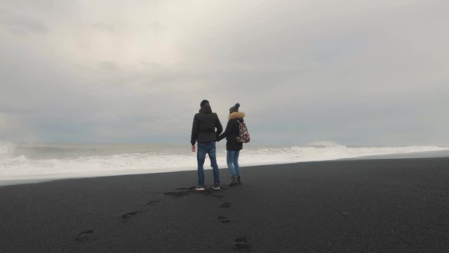Young hipster couple running on volcanic black sand beach in Iceland and having fun at sunset, slow motion. Reynisfjara Beach, Reynisdrangar troll toes mountains, Vik. Iceland in winter