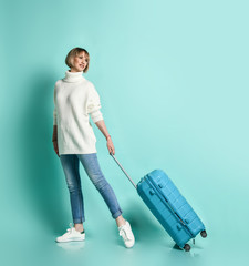 Blonde girl in white sweater, jeans and sneakers. She is smiling posing with suitcase luggage standing sideways on blue 