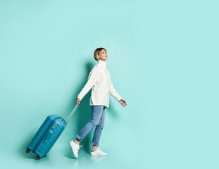 Blonde girl in white sweater, jeans and sneakers. She is smiling posing with suitcase luggage standing sideways on blue 