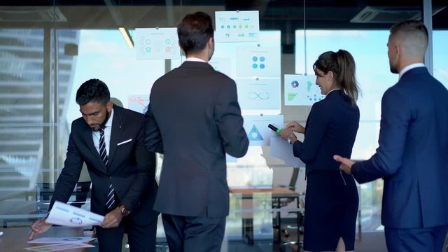 Multicultural group of corporate workers analyzing monetary gain of company capital standing near glass stand with paper reports on it and searching negotiations for exchange experience