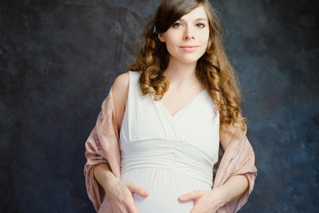 Fototapeta na wymiar Caucasian pregnant young woman on dark backdrop. Girl with wavy hair wearing white dress. Maternity, femininity concept.