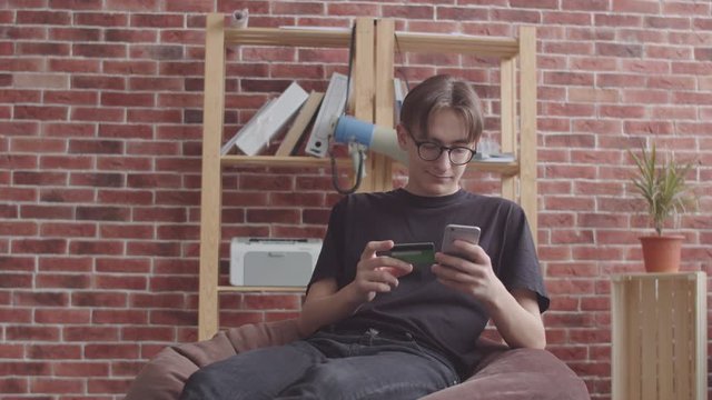 A thin guy sits in a bag chair and pays for a purchase in an online store with a Bank card. A hipster with a nose piercing sits in an office chair in a bag and enters credit card details into the