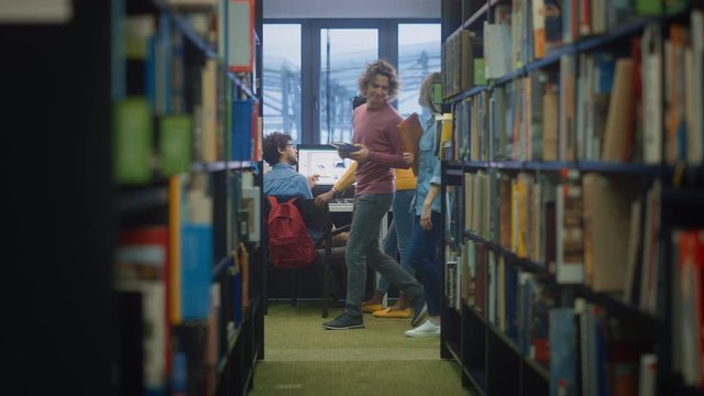 University Library: Boy Uses Personal Computer at His Desk, Talks with Girl Classmate who Explains, Helps Him with Class Assignment. Focused Students Study Together. Shot Between Rows of Bookshelves