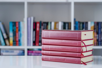Pile of red books literature stack on the white table in front of the bookshelf in background at home in day front view