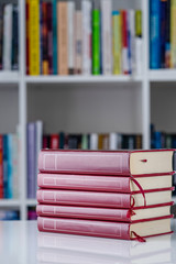 Pile of red books literature stack on the white table in front of the bookshelf in background at home in day front view