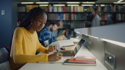 University Library: Gifted Black Girl uses Laptop, Writes Notes for the Paper, Essay, Study for Class Assignment. Diverse Multi-Ethnic Group of Students Learning, Studying for Exams, Talk in College - Powered by Adobe