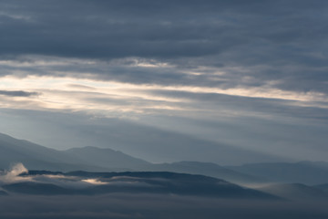 Fototapeta na wymiar clouds and rays of dawn in mountain valley, soft light in early morning, meditation in nature. Gentle hills in bluish haze, silhouettes of mountains