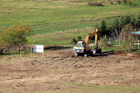 Freshly flattened and cleaned local construction site with single excavator parked after work surrounded with partially dry grass and small trees