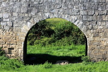 Massive old stone arch under stone bridge allowing access to local field surrounded with uncut grass and small plants