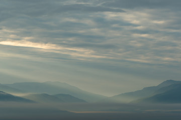 clouds and rays of dawn in mountain valley, soft light in early morning, meditation in nature. Gentle hills in bluish haze, silhouettes of mountains