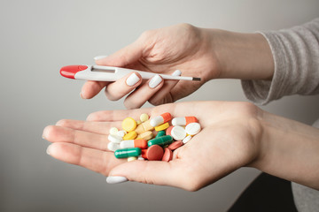 A handful of various pills with a digital thermometer in a woman's hands. Medications against the COVID-19 and other conditions and diseases on the grey background. Medicine.