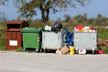 Various large metal and plastic trash containers for ecologically sorting garbage for recycling from glass and plastic to paper and fabric filled with garbage next to paved road surrounded with tall u