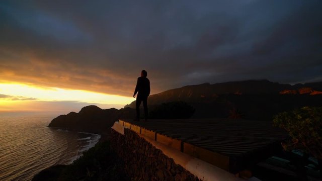 Man Standing At Cliff Edge Overlooking Sunset