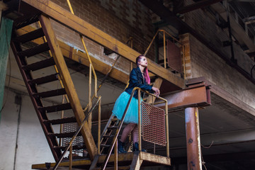 Portrait of a young girl with pink hair standing on the rusty stairs inside of collapsed building surrounded by ruins