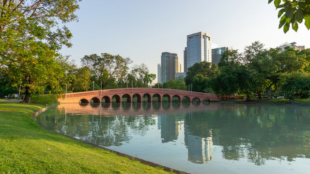 Beautiful garden in Chatuchak park Bankok Thailand, orange arch curve concrete brigeon a lake, building and greenery trees on background under blue sky in good care maintenance landscaped in a park