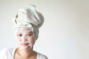 Asian Young woman with delighted expression, applies natural beauty product mask sheet on face, uncloges pores, has charming smile, looks aside, has wrapped towel on head, has naked body, healthy skin