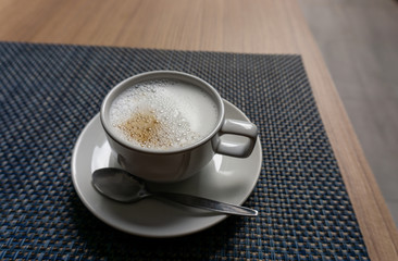 A white cup hot capuccino with white bubble mug, stanless spoon on ceramic plate on brown wooden table