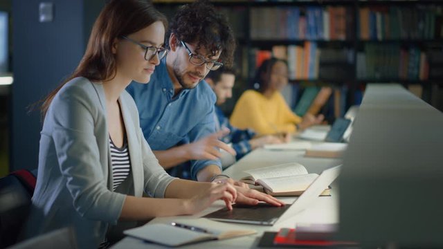 University Library: Talented Caucasian Girl uses Laptop, Smart Helpful Classmate Explains and Advices Her with Class Assignment. Happy Diverse Students Talking, Learning, Studying Together for Exams