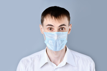 Man in a medical mask.   Surprised, scared man in a medical mask  in a white shirt looking at camera , on a gray background. Chinese Coronavirus , Covid-19