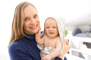 Cheerful new mom holding and hugging baby girl wrapped in hooded bathrobe. Young woman nursing six month daughter at home. Motherhood concept