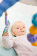 Cute adorable baby girl playing with hanging rattle. Six month little child lying in bed. Childhood or baby care concept