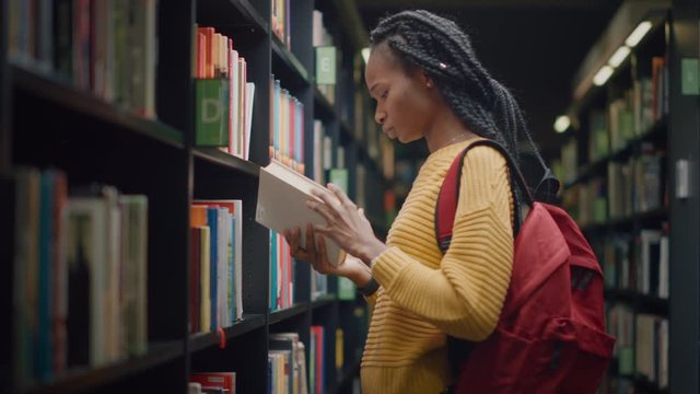University Library: Portrait of Gifted Beautiful Black Girl Walking Between Rows of Bookshelves Using Smartphone Searching for the Right Book Title, Finds and Picks one for Class Assignment