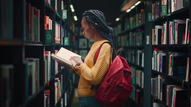 University Library: Portrait of Gifted Beautiful Black Girl Walking Between Rows of Bookshelves Using Smartphone Searching for the Right Book Title, Finds and Picks one for Class Assignment