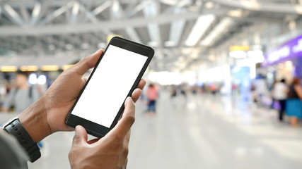 Smart man's hands holding a cropped black smartphone with white blank screen over crowded international airport as blurred background.