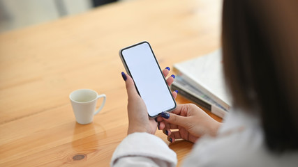 Cropped image of young creative woman's hands holding a smartphone with empty screen while sitting at the working desk over comfortable living room as background.