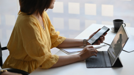 Cropped image of beautiful woman working as secretary holding a white blank screen smartphone and stylus pen while sitting in front her computer tablet with keyboard case over office as background.