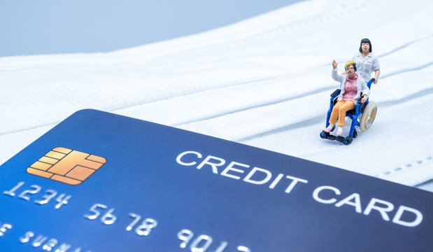 Miniature doctor send old women in wheelchair back home during crisis covid-19, with a credit card and mask on gray background. Medical, finance and healthcare concept.