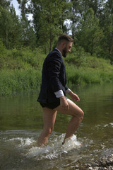 Bearded young businessman in black classic suit jacket, boxer underwear run ankle-deep in water of narrow forest river