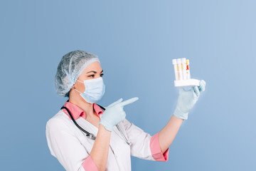 Young girl doctor in a white uniform, in gloves, in a mask holding a tray in front of him with test tubes for analysis on a blue background