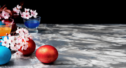 Painted Easter eggs on a gray background, flowering branches of cherries next to the table. Closeup, copy space for text. Easter preparations