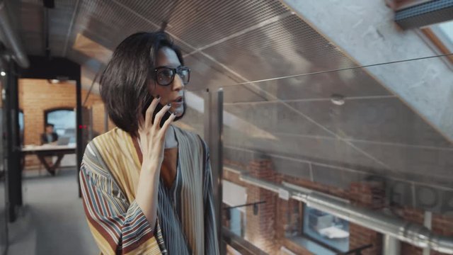 Waist up shot of brunette businesswoman in casual outfit and glasses speaking on mobile phone while walking along glass wall in office
