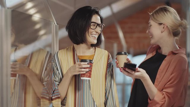 Two beautiful blonde and brunette businesswomen in casualwear watching social media post on smartphone, chatting and laughing while having coffee break in office
