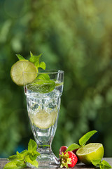 in a tall glass a refreshing drink with the addition of mint, lime and ice, against a background of greenery, next to strawberries and mint leaves