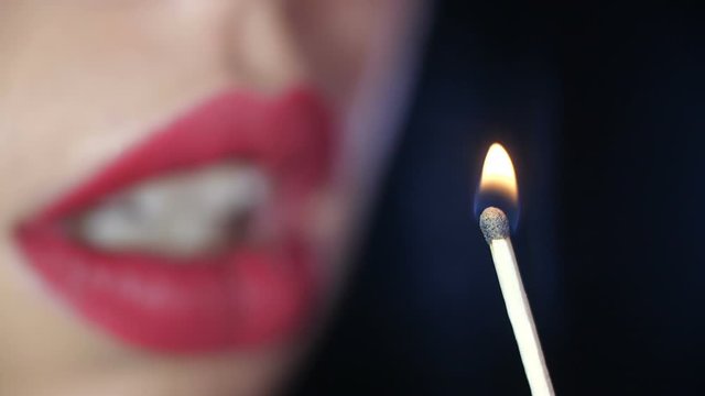 Detail close-up macro shot of the unfocused lips of a girl painted with a matte burgundy lipstick  blowing out the fire of a burning match on black studio background isolated. 4k slow motion 50 fps