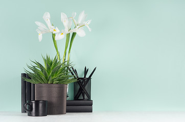 Cozy home workplace with black stationery, books, coffee cup, green aloe, fresh spring white iris bouquet in trend green mint menthe color interior and white wood table.